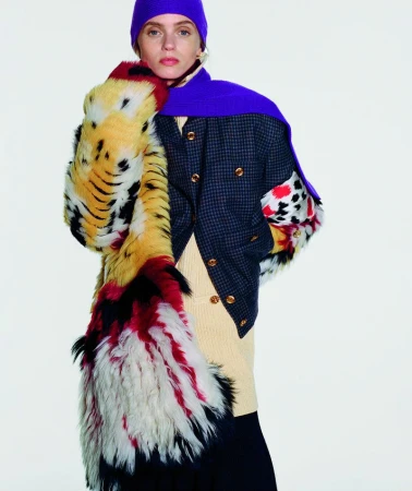 Model Abbey Lee poses in a purple knit headpiece, patterned faux fur coat, and plaid jacket against a white studio background.