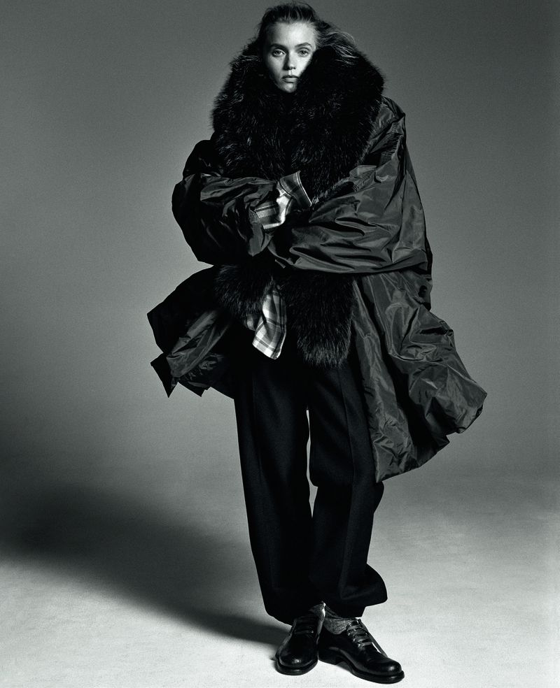 Model Abbey Lee posing in an oversized dark coat with a voluminous fur collar and wide-leg trousers on a studio backdrop.