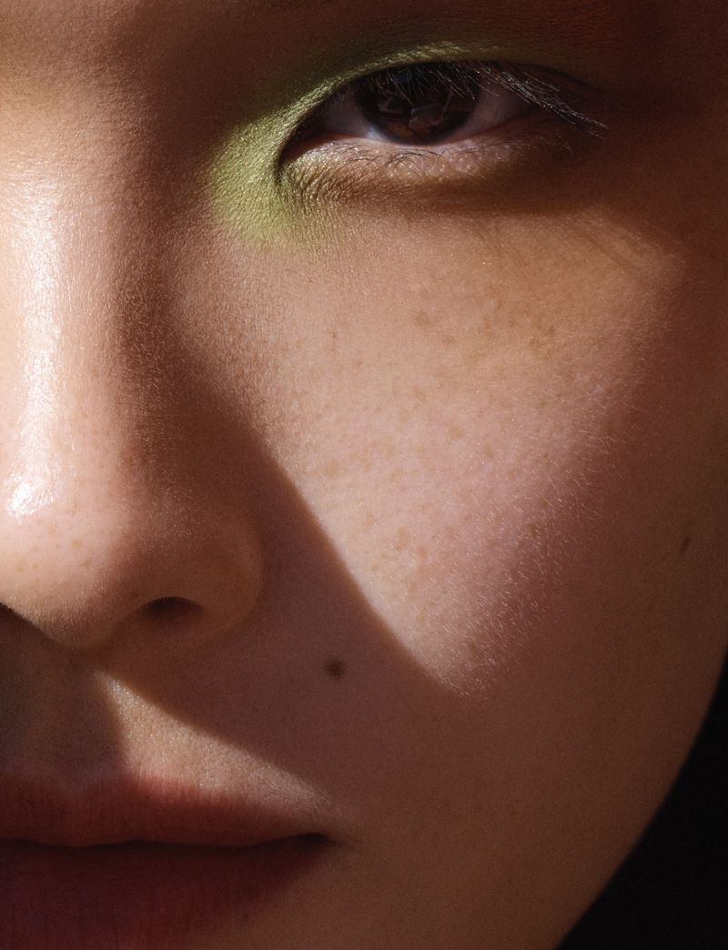 Macro close-up portrait of model Xie Chaoyu with green eyeshadow and freckles under diagonal hard light.