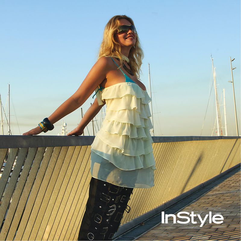 A blonde woman in a ruffled cream top and black buckled pants leans against a marina railing on a sunny day.