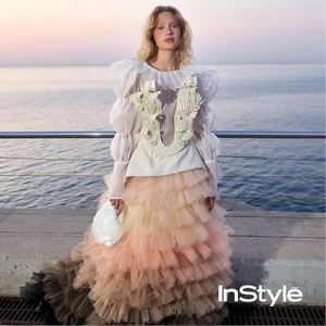 Fashion model posing by the sea at sunset wearing an ornate white blouse with a corset and a voluminous tiered tulle ombre skirt.