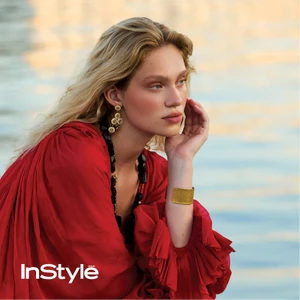 Woman wearing a vibrant red ruffled dress, gold drop earrings, and a gold cuff bracelet posed against water.