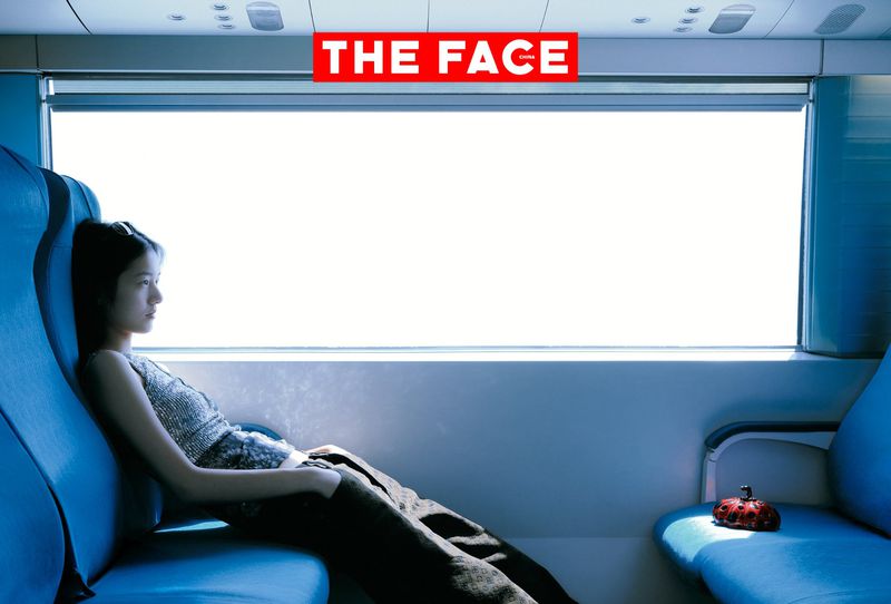 A young woman in a grey top and dark pants leans back in a blue train seat next to a bright window.