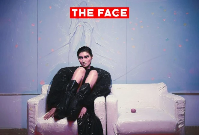 Model wearing a voluminous black dress and tall black boots seated on a white textured armchair against a painted pastel wall.