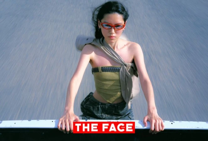 East Asian model in orange sunglasses and an olive tube top leaning over the back of a moving vehicle on a paved road.