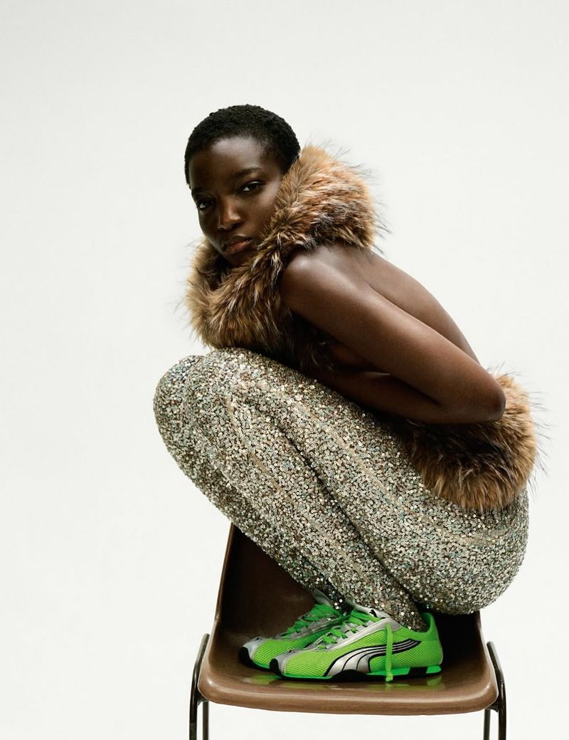 A solitary black model crouching on a chair with a fur-trimmed vest, sparkly pants, and lime green cleats against a white backdrop.