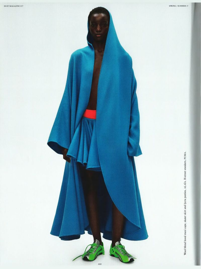 Fashion editorial shot of a Black female model wearing a large turquoise hooded coat and skirt with lime green athletic shoes against white.
