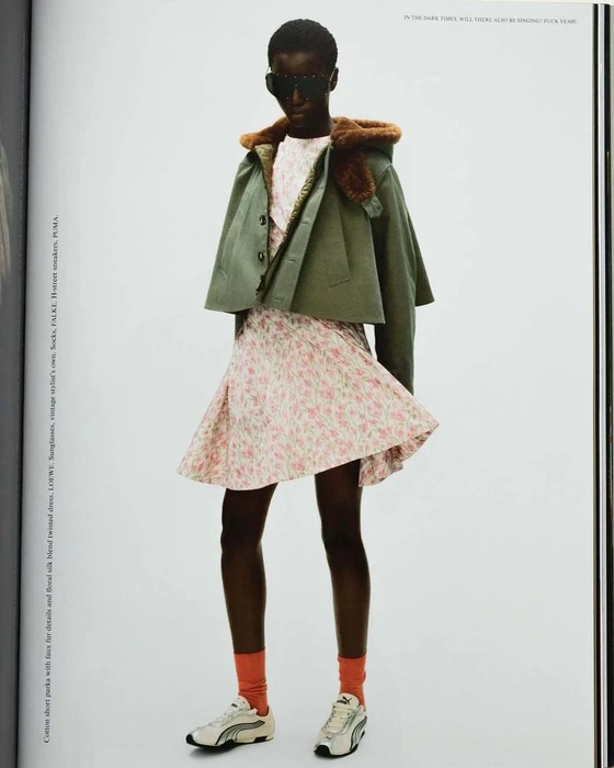 Model wearing dark sunglasses, cropped green jacket, patterned dress, and sneakers against a white background.