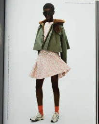 Model wearing dark sunglasses, cropped green jacket, patterned dress, and sneakers against a white background.