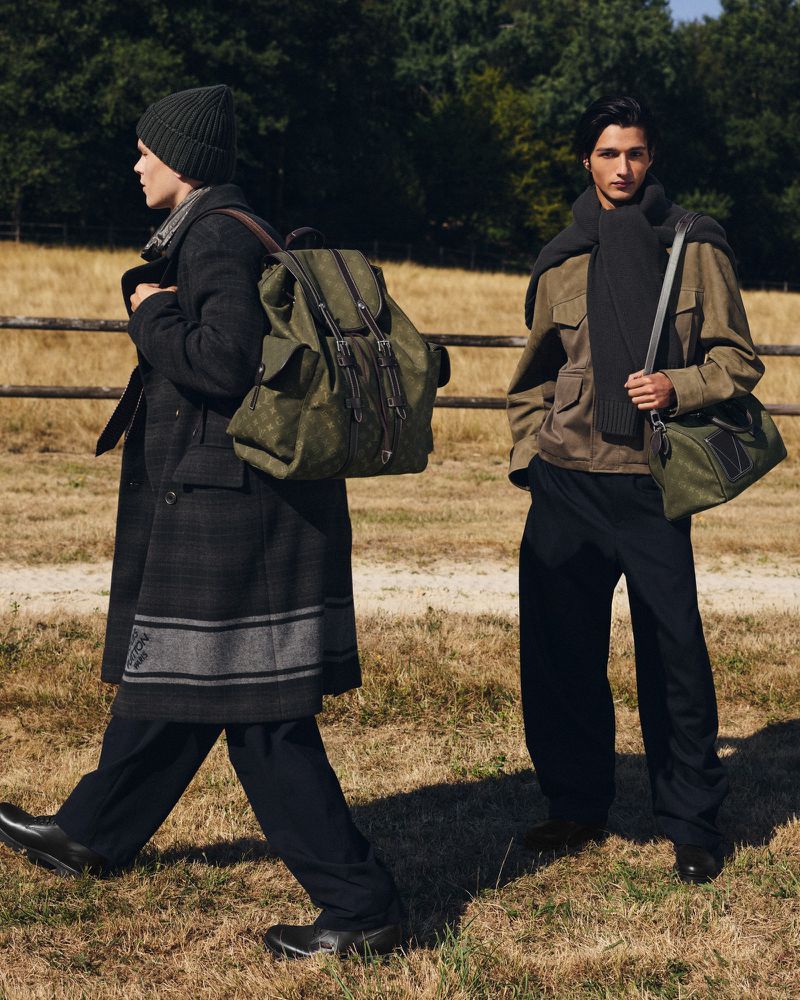 Two male models in designer outerwear with olive green monogram luggage in an open grassy field with a wooden fence.