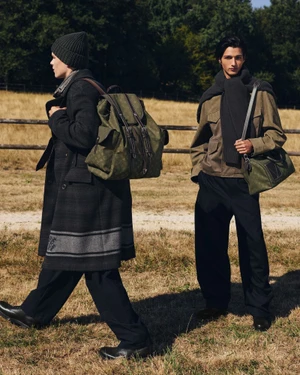 Two male models in designer outerwear with olive green monogram luggage in an open grassy field with a wooden fence.