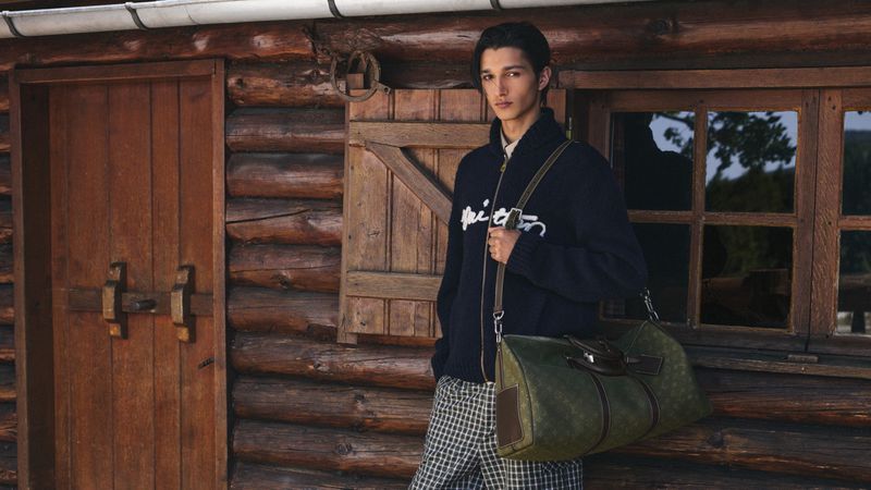 Model Rubuen Bilan-Carroll wearing a navy zip cardigan and holding an olive green duffle bag outside a log cabin.