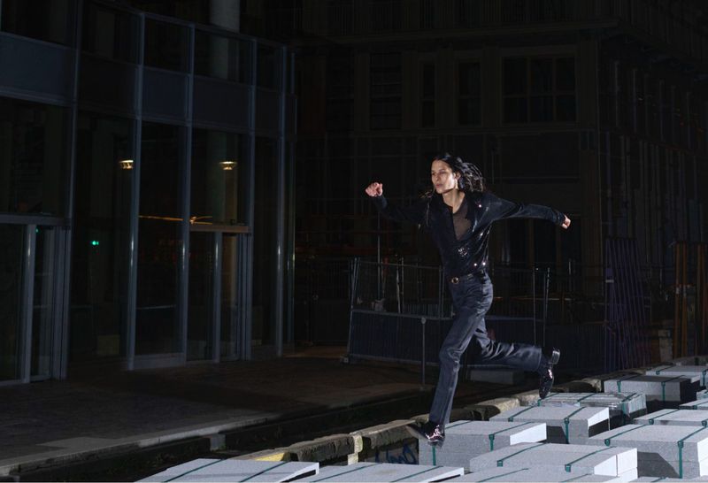 Adult woman runs across blocks in an urban setting at night. She is wearing a black jacket and jeans in a low-angle outdoor shot.