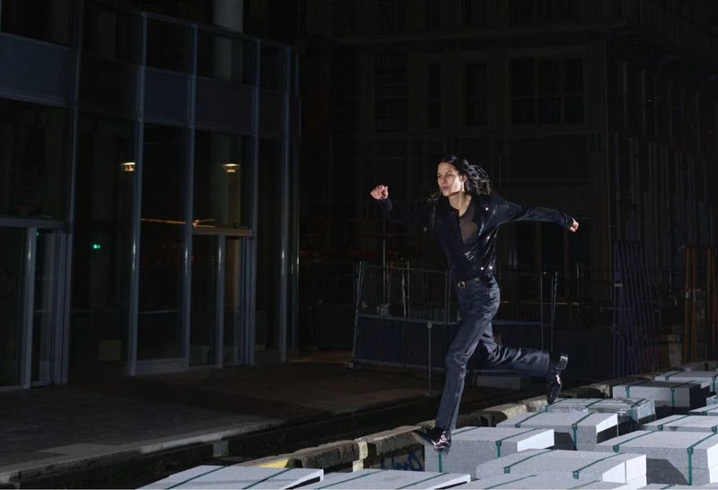 Adult woman runs across blocks in an urban setting at night. She is wearing a black jacket and jeans in a low-angle outdoor shot.