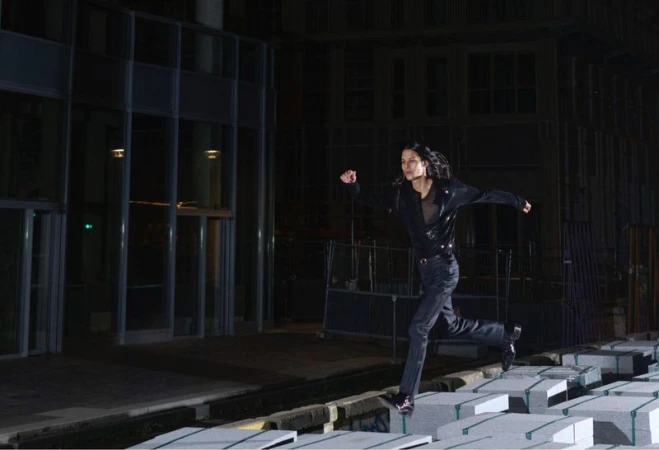 Adult woman runs across blocks in an urban setting at night. She is wearing a black jacket and jeans in a low-angle outdoor shot.