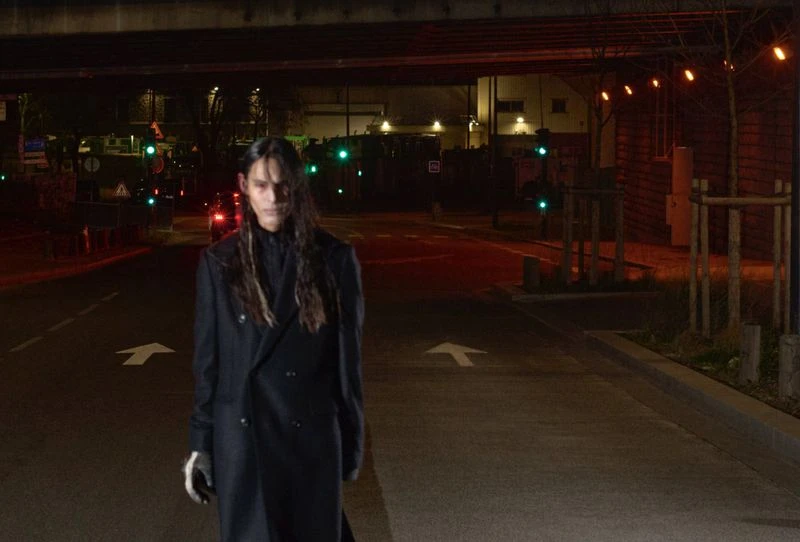 Person in a black coat and gloves stands on a street under a bridge at night with traffic signals and city buildings in the background.