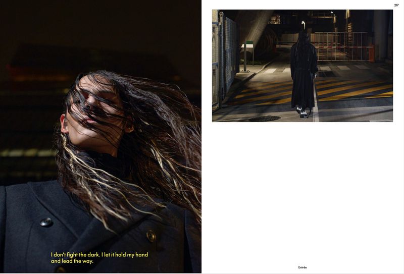 Woman with windswept highlighted hair, wearing a dark coat in dark lighting. Full length view of person in black coat on city street.