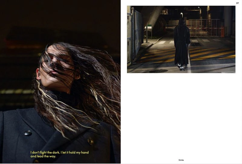 Woman with windswept highlighted hair, wearing a dark coat in dark lighting. Full length view of person in black coat on city street.