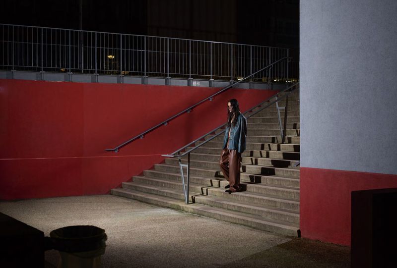 A woman walks down stone stairs in an urban setting, wearing a blue jacket and brown pants. Red painted wall in the background at night.