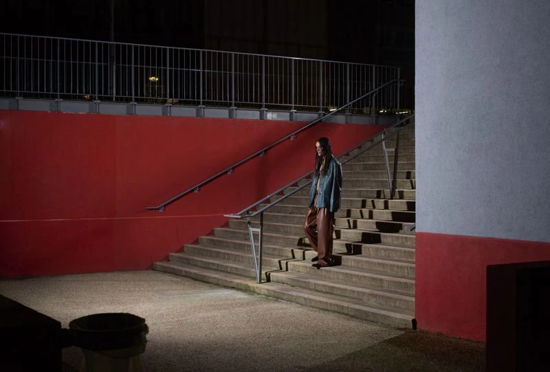 A woman walks down stone stairs in an urban setting, wearing a blue jacket and brown pants. Red painted wall in the background at night.