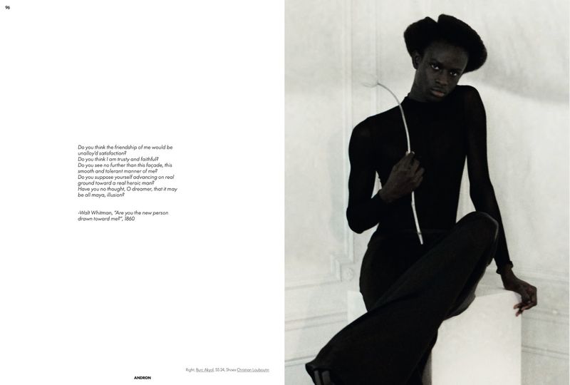 A slender African-descent adult sits, wearing black clothing and hat, posed against a light-colored interior wall in a fashion-style shot.