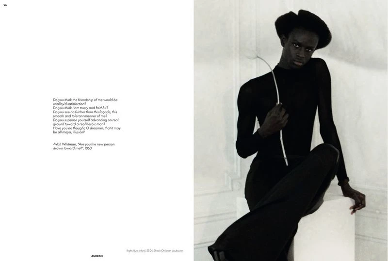 A slender African-descent adult sits, wearing black clothing and hat, posed against a light-colored interior wall in a fashion-style shot.