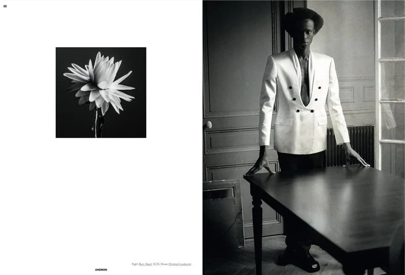 Monochrome fashion shot of a Black male model in a white jacket at a table, alongside a flower image on a magazine spread.