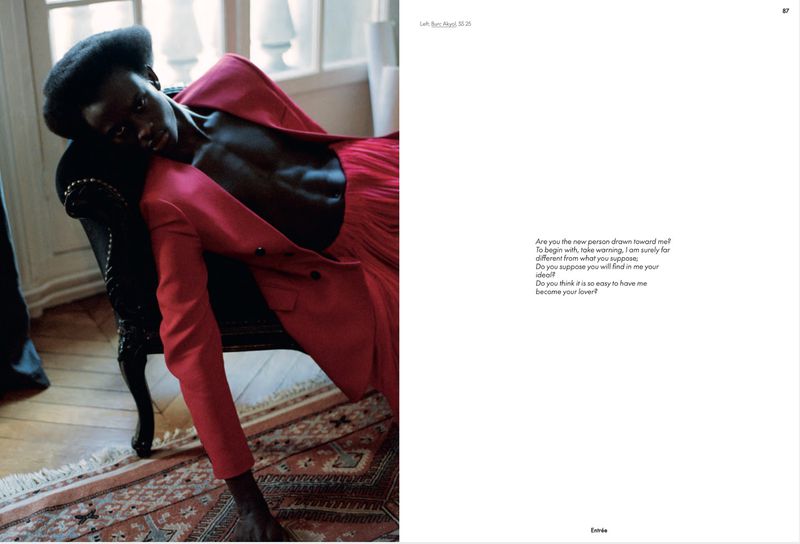 Dark-skinned model in a red suit reclines on a black chair in a natural light studio with a book featuring poetic text.