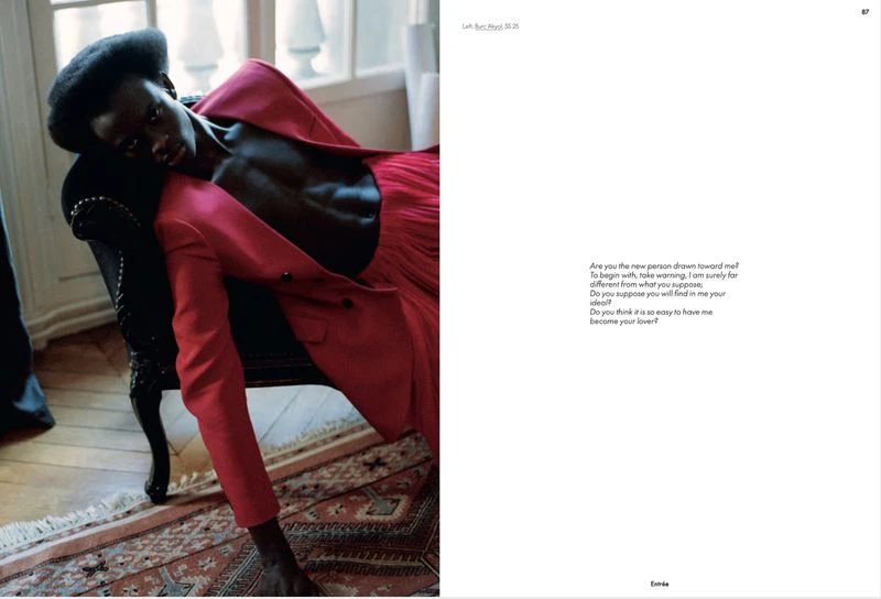 Dark-skinned model in a red suit reclines on a black chair in a natural light studio with a book featuring poetic text.
