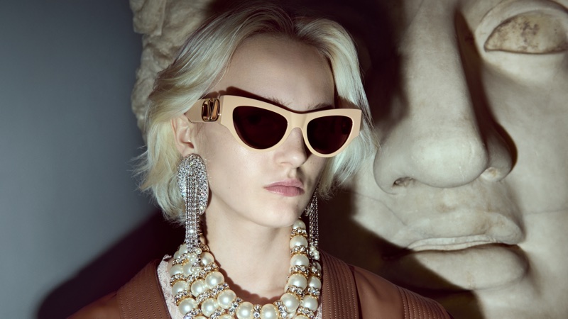 Blonde model wearing beige cat-eye Valentino sunglasses, ornate crystal earrings, and layered pearl necklaces beside a marble sculpture.
