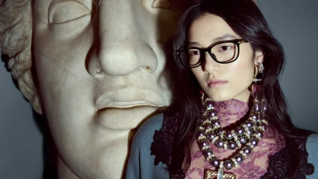 Model wearing eyeglasses and beaded necklace against a large classical stone sculpture backdrop.