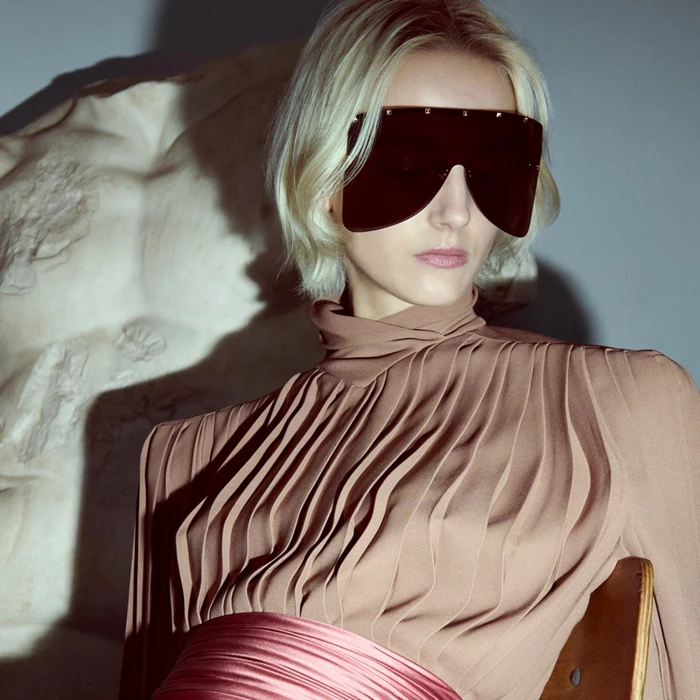 Model with blonde hair wearing dark shield sunglasses and a pleated taupe top against a sculptural background.