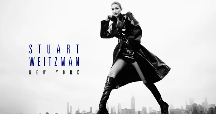 Gigi Hadid in a black patent trench coat and tall boots against an urban skyline backdrop for Stuart Weitzman.