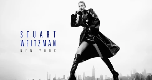 Gigi Hadid in a black patent trench coat and tall boots against an urban skyline backdrop for Stuart Weitzman.