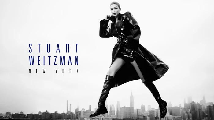 Gigi Hadid wearing a black patent leather coat and tall boots leaping against a cityscape background for Stuart Weitzman.