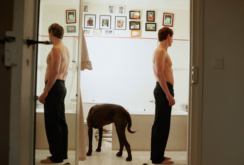 Nude man and brown dog stand in a white-walled bathroom with a bathtub and mirror reflecting the man from the back.