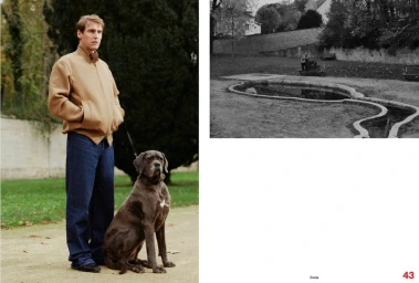 Man wearing a camel jacket and blue jeans standing outdoors next to a large grey dog on a leash