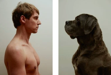 Profile portrait of a shirtless man facing a large dark dog in a studio setting.