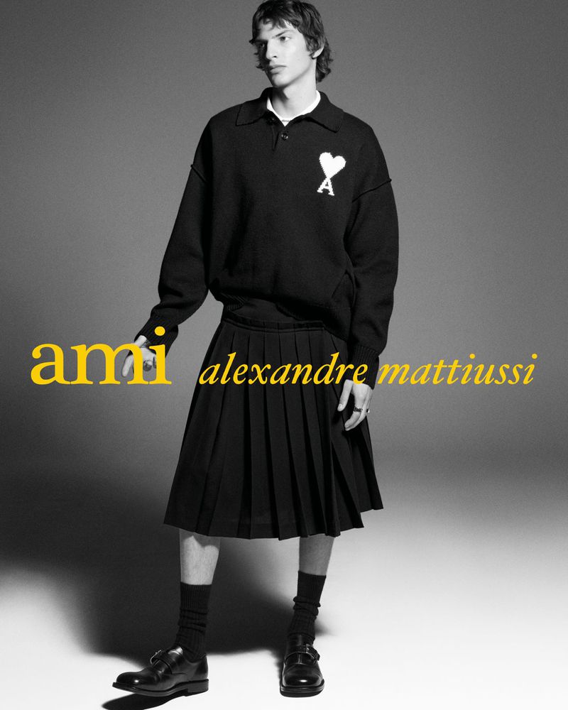 Male model wearing a black polo sweater and black pleated kilt skirt against a grey studio background for Ami.