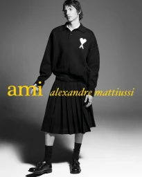 Male model wearing a black polo sweater and black pleated kilt skirt against a grey studio background for Ami.
