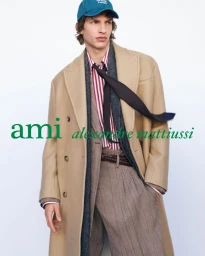 Male model wearing an oversized camel double-breasted coat, striped shirt, windblown black tie, and teal cap against a white background.