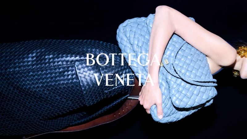 Model holding a light blue woven leather Bottega Veneta handbag beside a black woven tote against a black backdrop.