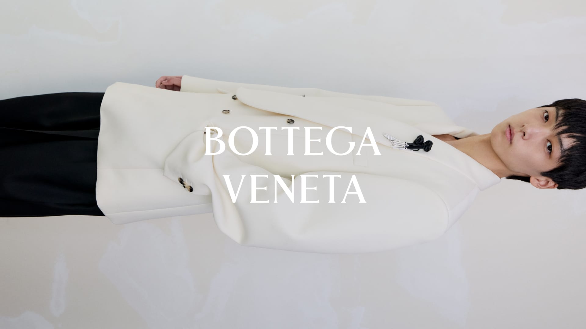 East Asian man wearing an off-white double-breasted blazer and black trousers in a studio setting with Bottega Veneta typography.