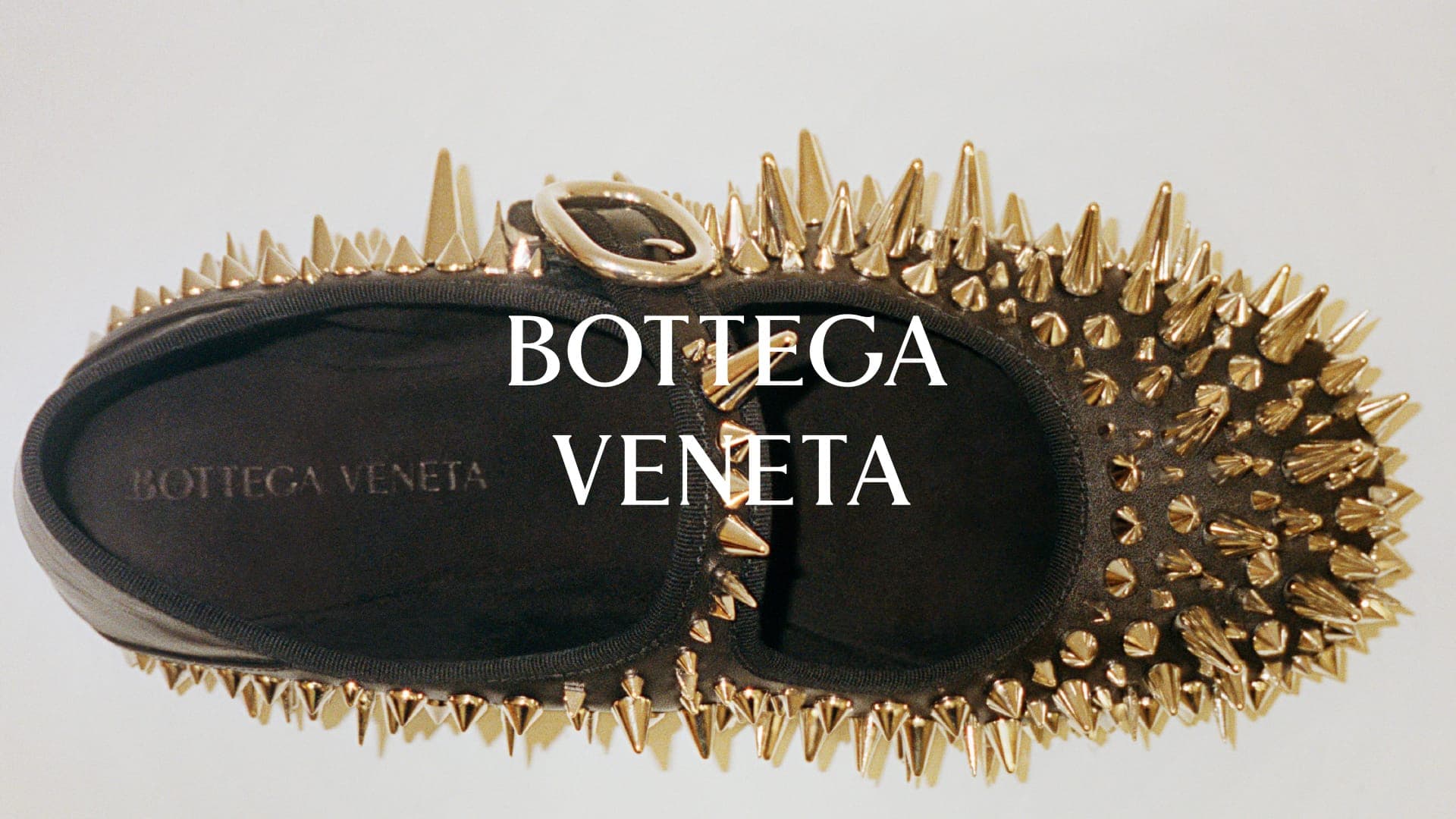 Top-down view of a black Bottega Veneta flat shoe covered with sharp metallic gold spikes and an instep buckle strap on a white background.