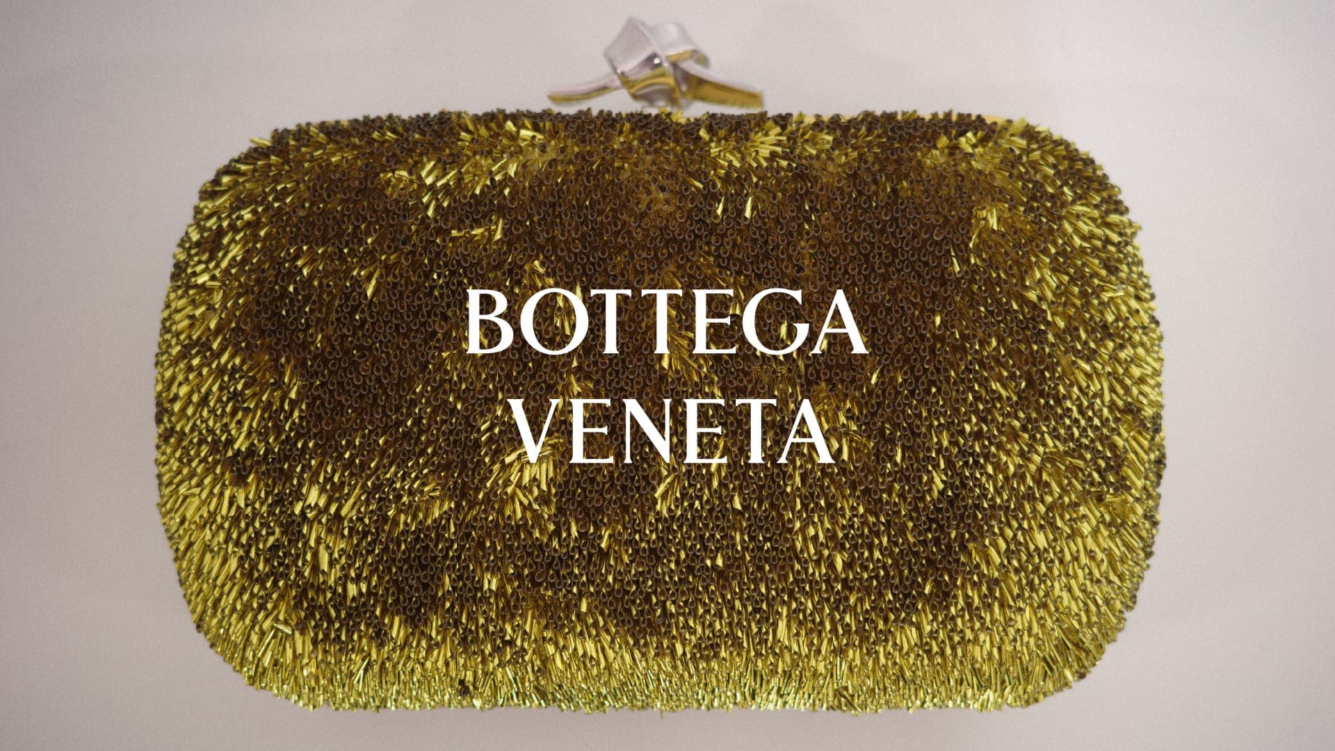 Bottega Veneta metallic gold textured fringe clutch bag with a sculptural silver knot clasp centered on a pale seamless backdrop.