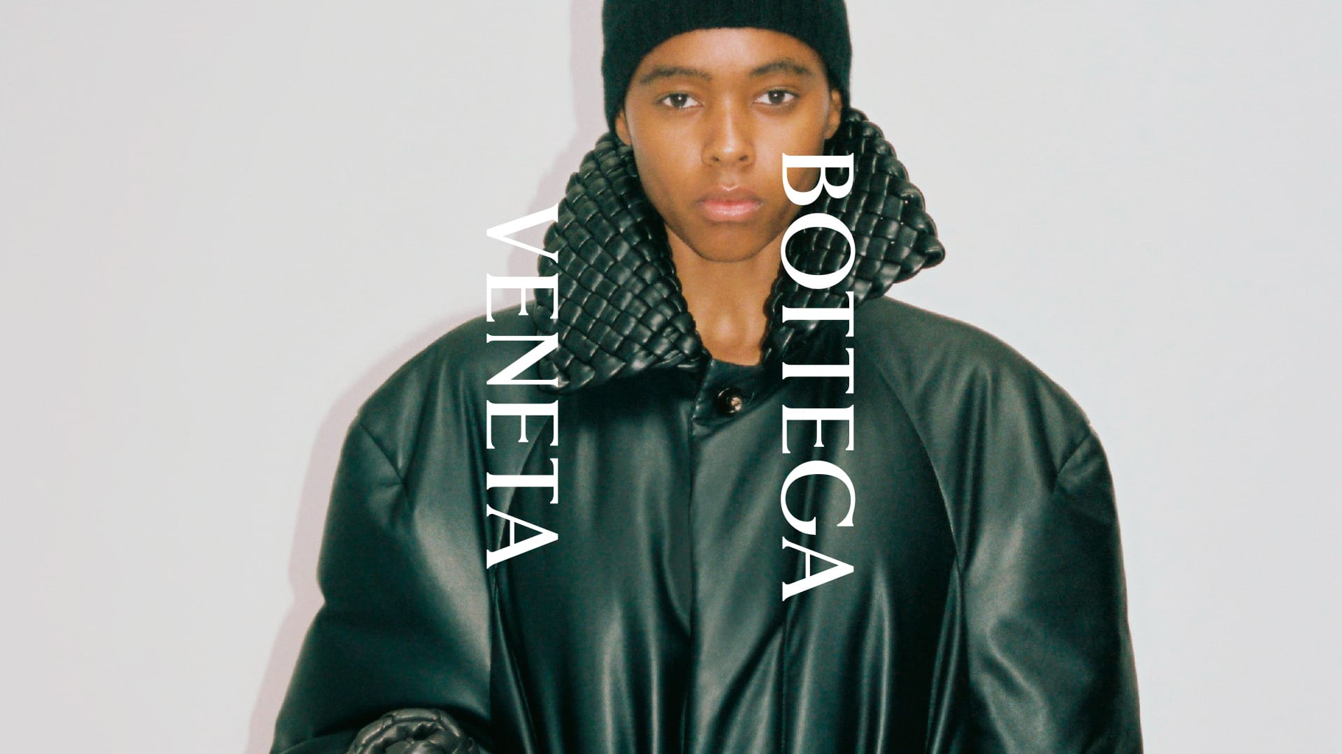 Model wearing an oversized dark green leather coat with a woven intrecciato collar and black beanie against a white studio background.