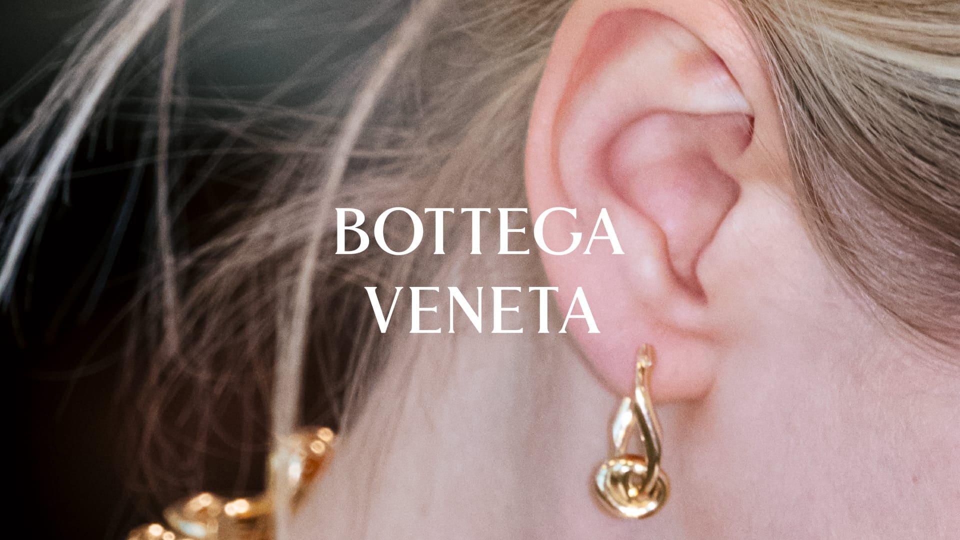 Close-up of a model's ear wearing a sculptural gold drop earring with the white Bottega Veneta logo centered.