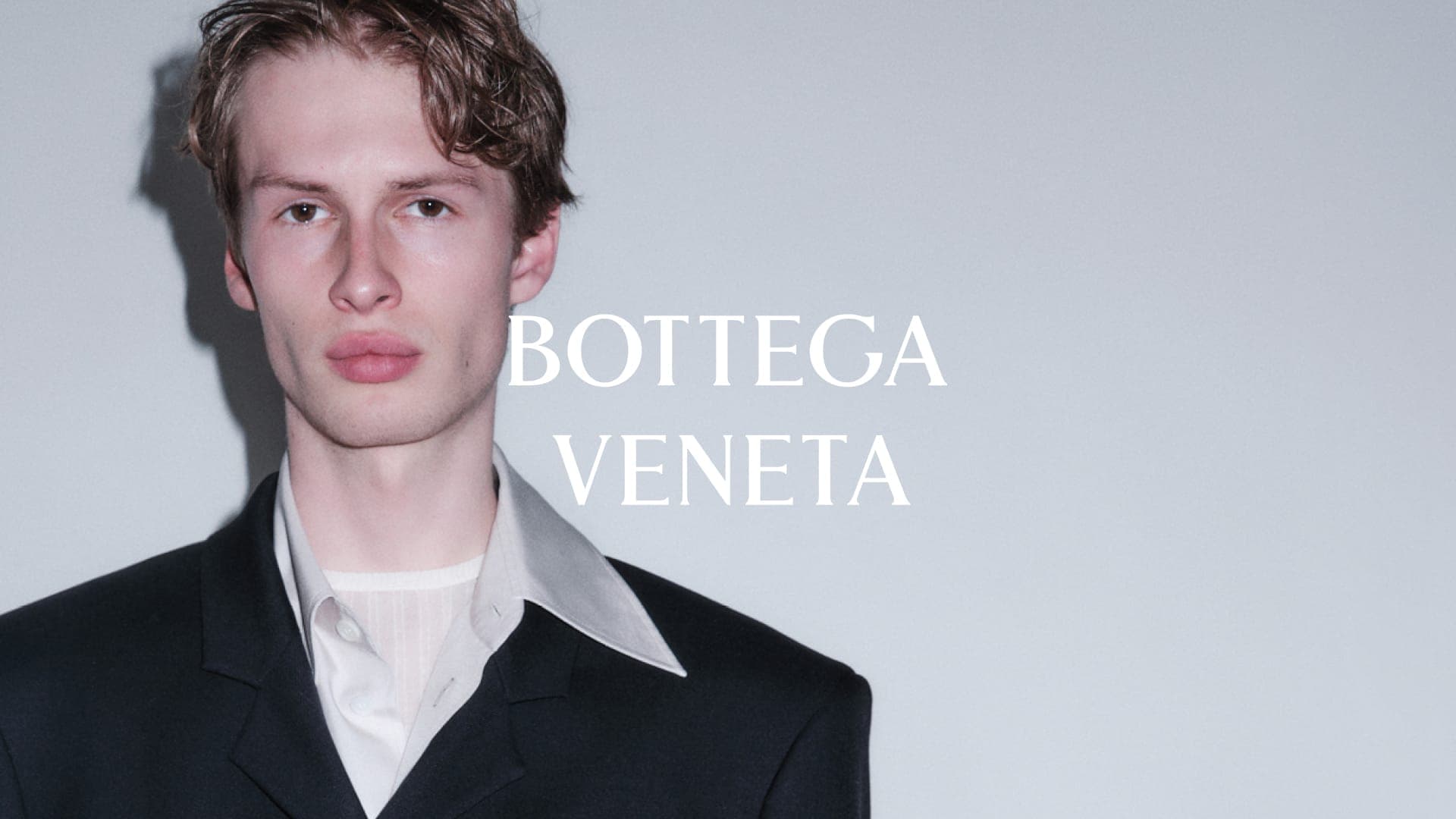Male model wearing a black tailored blazer and white collared shirt against a light grey wall with the Bottega Veneta logo.