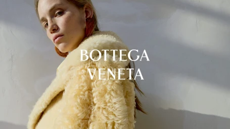 Model wearing a pale yellow textured shearling coat with Bottega Veneta logo overlay against a sunlit wall.