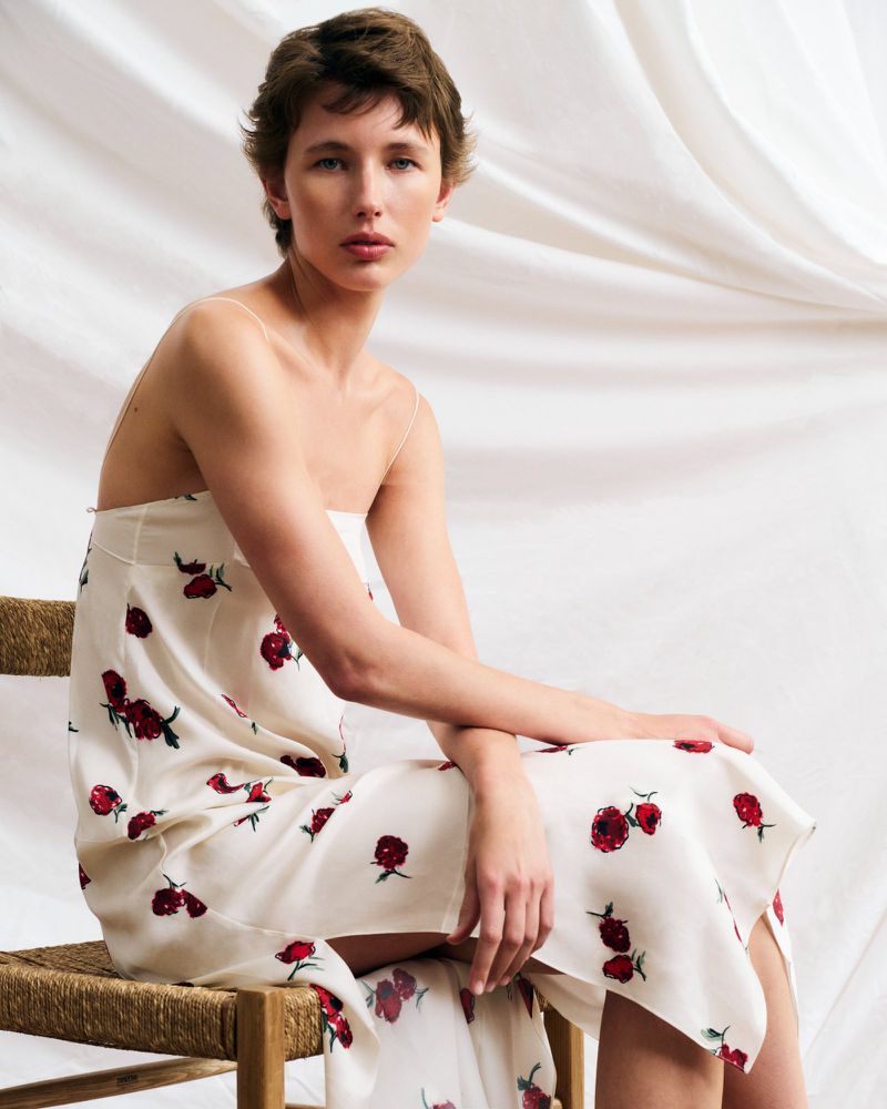 Adult woman in white spaghetti strap dress with red floral pattern seated on woven chair in front of white curtain backdrop.